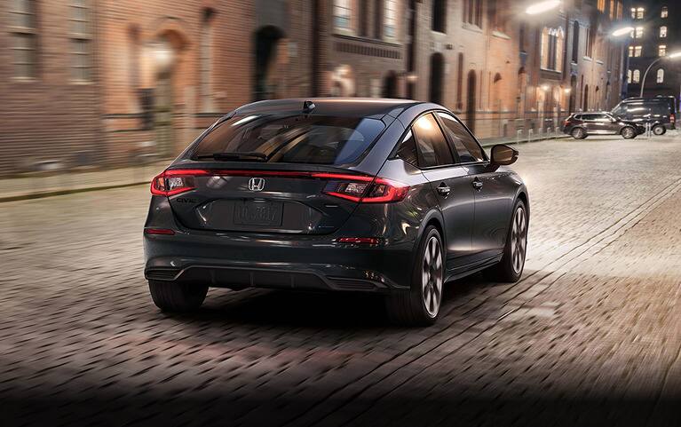 2027 Honda Civic Hatchback Sport Touring Hybrid in Meteorite Gray Metallic back driving on a cobblestone city street (Compact Hatchback).