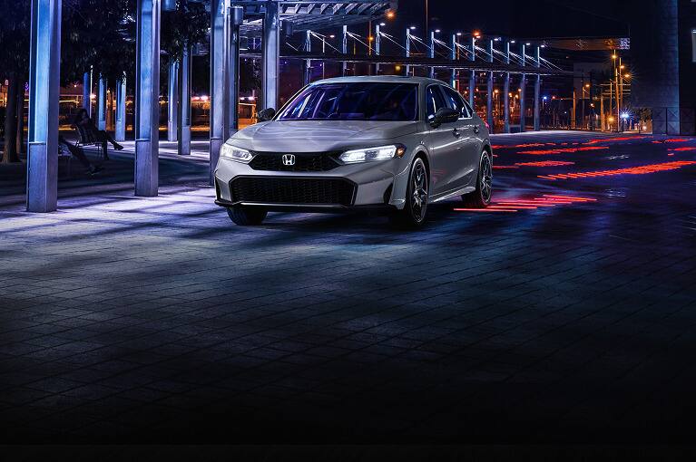 2027 Honda Civic Sedan Sport Hybrid in Urban Grey Pearl front parked in a modern urban plaza at night (Compact Sedan).
